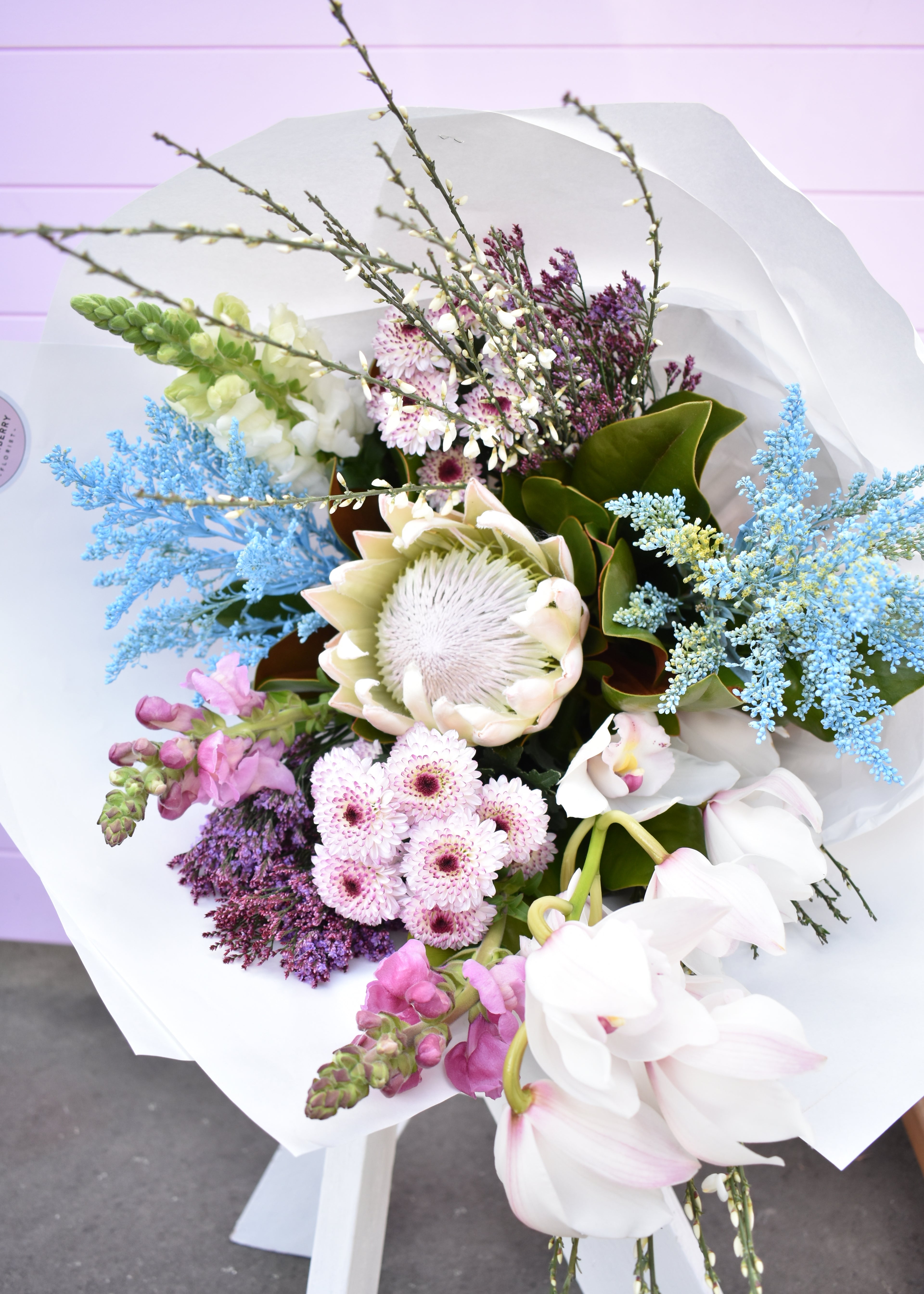 Luxurious Premium florist bouquet created in white, pale blue and mauve colours. Featuring a white king protea, orchid and chrysanthemums. Presented in white paper wrap.